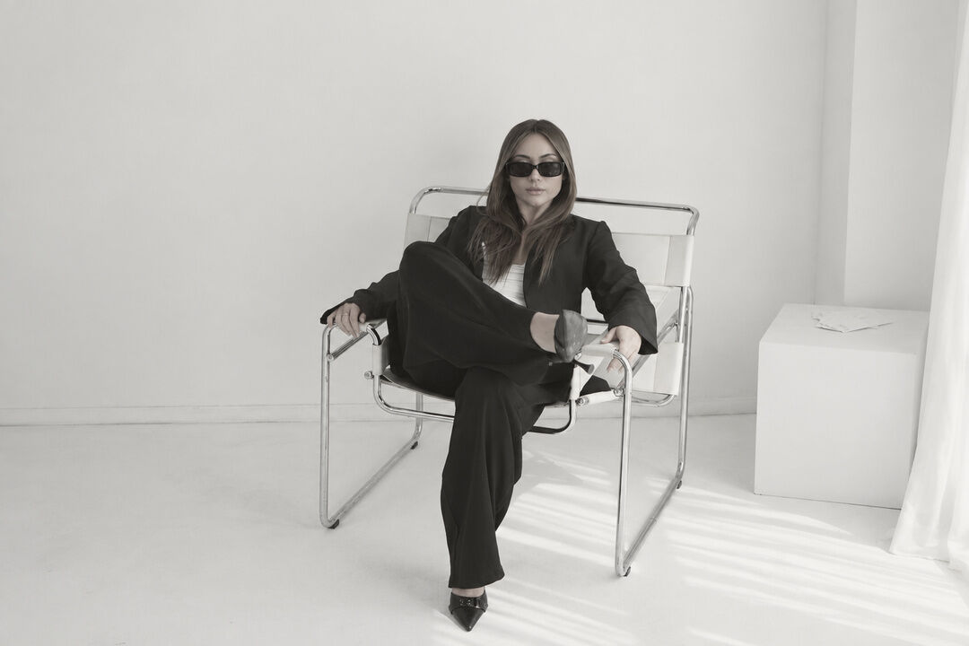 Pia sentada en una silla blanca con anteojos y saco negro